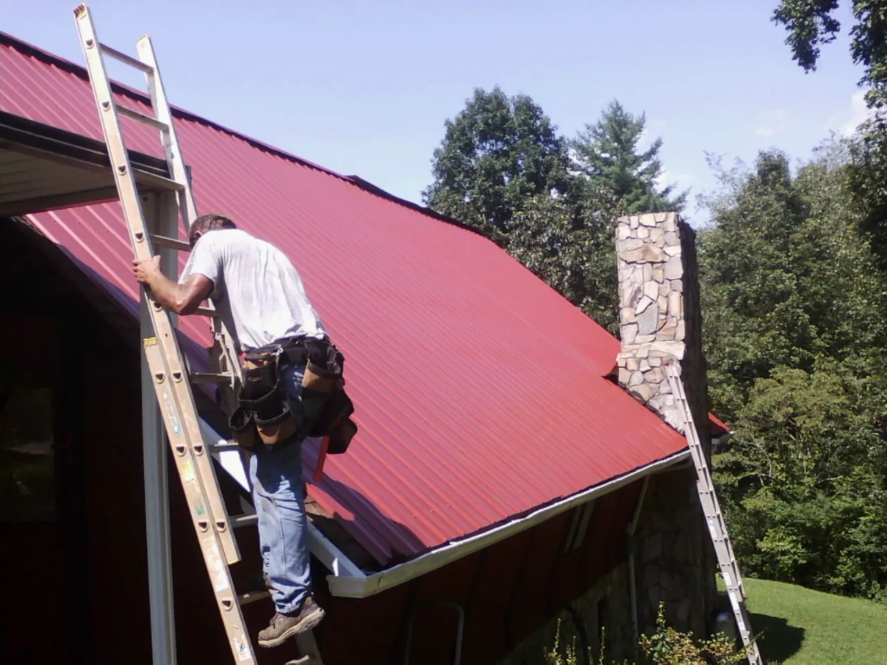 Roofer on a ladder beside a red metal roof and stone chimney — Soffit & Fascia Repair by Roofing Brothers in Wynantskill